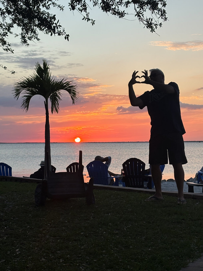 A silhouette of a man with a heart symbol, including wings, stands in front of a sunset on the harbor. A palm tree and a beach goer is in the foreground, with an oak above.