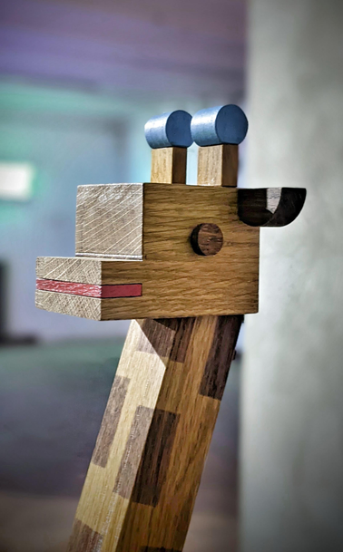 Ein eckiges Kunstwerk in Form eines Giraffenkopf und -Halses aus Holz vor unscharfem Hintergrund in grau, verwaschenem grün und milchigen lila. Trotz der Eckigkeit der Holzmodule sieht das Tier sehr süß aus, wie ein Kinderspielzeug, was durch das Auge, die Ohren und die Enden der Hornzapfen auf dem Kopf  ausgelöst wird: rund geformt und andersfarbig (dunkelbraun, fast schwarz und blau), der Kontrast verstärkt die Wirkung.