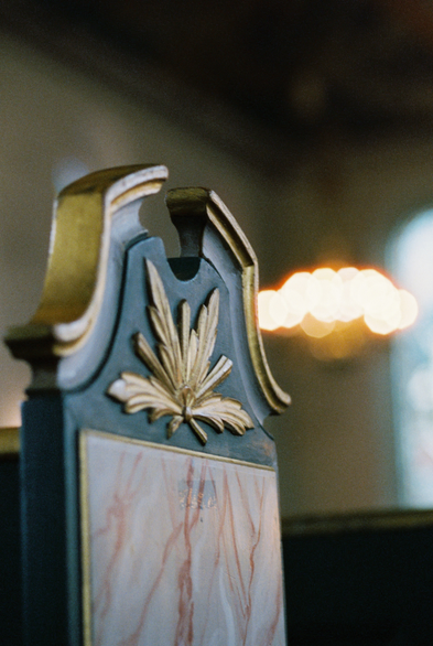 Close-up photo of the side of a wooden pew in a church. The pew is painted green and gold, and it is decorated with carved golden leaves and a marbled inset on the end. In the blurry background, a chandelier and a tall but barely-visible window work together to bring some light to the scene.