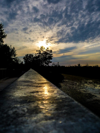 The evening sun begins to set behind the clouds and the silhouette of distant trees.  A pond is seen to the right and the camera sits on a metal railing and the sun can be seen reflecting off the shiny surface.