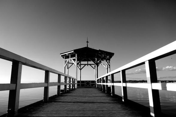 Picture showing a wooden structure as planks to a pavilion at the waterfront of a lake. Water around and blue sky is the setting. No humans or animals are seen.