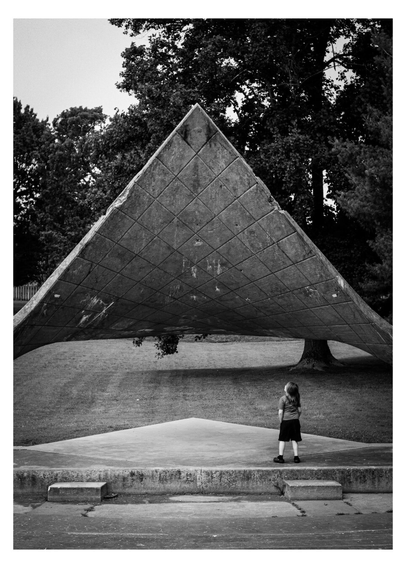 Black and white photo of an odd triangular outdoor auditorium in a park. A person with long hair is standing underneath it with their hands in their pockets looking up at it.