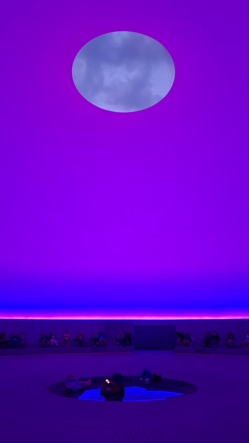 Same sunrise, different angles and colours and times. Same Alt Text follows for all photos. Short version: colours and effects in a skyspace dome. Fuller description: What looks like the Earth or the moon in a coloured ‘sky’ above us. The ‘planet’ is really an aperture in the room of the dome we’re standing inside. We are seeing the sky changing colour at dawn, clouds making shapes as they move – a midsummer sunrise dawning between 3am and 4 am. This is inside the underground Skyspace dome ‘As Seen Below’, designed by artist James Turrell. Part of ARoS Art Museum, Aarhus, Denmark. The Dome is mostly underground – 40 metres in diameter, 16 metres high. As people view the sunrise, vivid colours change on the concrete walls and ground of the vast circular room that has a special acoustic too. It is a hard-to-describe immersive experience. In the photos, I think it’s hard to know what you are looking at – it’s difficult to tell the ‘planet’ is a hole or that it’s high above us.