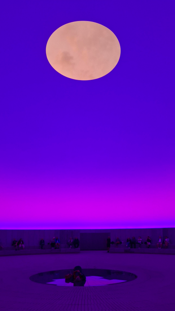 Same sunrise, different angles and colours and times. Same Alt Text follows for all photos. Short version: colours and effects in a skyspace dome. Fuller description: What looks like the Earth or the moon in a coloured ‘sky’ above us. The ‘planet’ is really an aperture in the room of the dome we’re standing inside. We are seeing the sky changing colour at dawn, clouds making shapes as they move – a midsummer sunrise dawning between 3am and 4 am. This is inside the underground Skyspace dome ‘As Seen Below’, designed by artist James Turrell. Part of ARoS Art Museum, Aarhus, Denmark. The Dome is mostly underground – 40 metres in diameter, 16 metres high. As people view the sunrise, vivid colours change on the concrete walls and ground of the vast circular room that has a special acoustic too. It is a hard-to-describe immersive experience. In the photos, I think it’s hard to know what you are looking at – it’s difficult to tell the ‘planet’ is a hole or that it’s high above us.