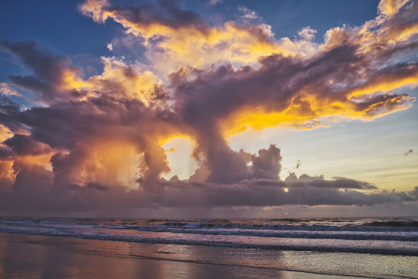 Serene beach sunrise with sun reflecting on wet sand and waves. The sky is filled with dramatic clouds, creating a peaceful, tranquil atmosphere.