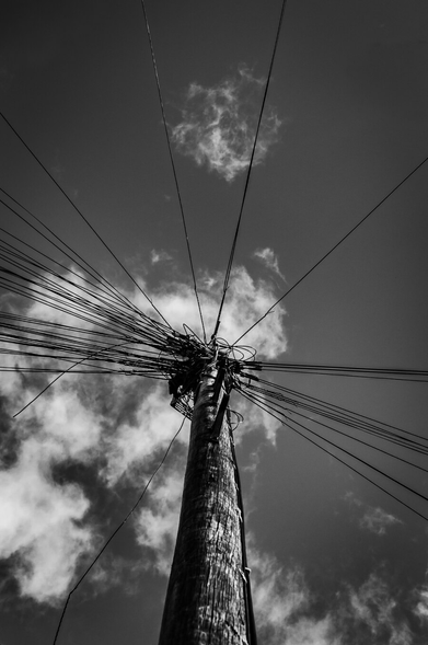 This is a black and white, low-angle shot looking up at a wooden utility pole. The pole itself is textured and appears weathered, with visible vertical grain and some signs of wear. It occupies the lower half of the frame, rising towards the top.
The most striking element is the dense tangle of black wires sprouting from the top of the pole, fanning out in numerous directions against the sky. The sky above is filled with dramatic, textured clouds, rendered in varying shades of gray.