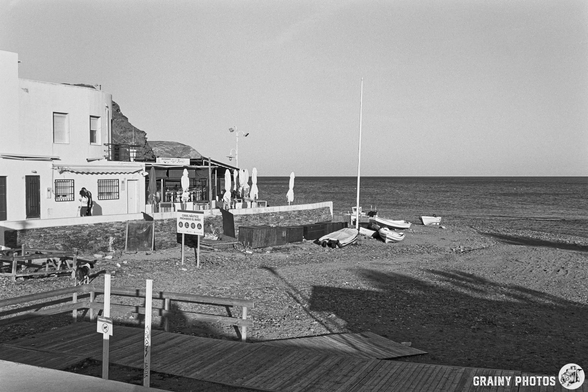A coastal scene featuring a beach with a wooden walkway, a small restaurant with outdoor seating, and boats moored nearby. The image is captured in black and white, highlighting the tranquil atmosphere and simplicity of the seaside setting.