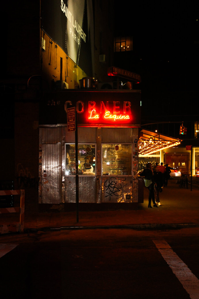 A high-contrast night photo of a New York street corner. A vibrant red neon sign reading "La Esquina" glows above a small, metallic sided storefront with graffiti and illuminated windows showing the interior. A Saint Laurent Paris billboard is visible on the upper dark brick wall. To the right, customers stand at the restaurant's walk up window under a brightly lit canopy.