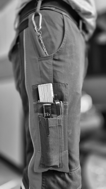 Ein rechteckiges Hochkantfoto in schwarzweiß von einer Person in Arbeitshose. Die Person wurde von der rechten Seite aufgenommen und ist nur im Bereich der Arbeitshose, also von etwa der Hüfte bis zum Knie, im Bild zu sehen. Zu sehen ist ein Karabinerhaken, der an einer Gürtelschlaufe befestigt ist. Daran verläuft eine Kette in die Hosentasche. Weiter unten ist eine Seitentaschen zu sehen, in denen sich ein Meterstab (Gliedermaßstab), eine kleine Stabtaschenleuchte, zwei Kugelschreiber und ein Bleistift befinden. All diese Gegenstände schauen nur zu einem Teil aus der Tasche. 
