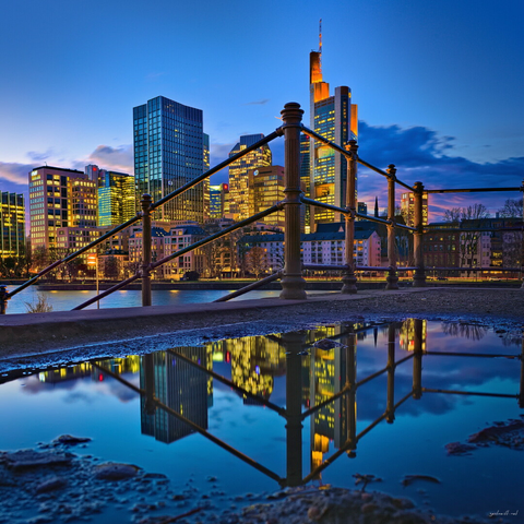 Evening city skyline reflected in a large puddle beside a riverside walkway. Illuminated glass skyscrapers glow gold and blue against a deep twilight sky, while metal railings in the foreground create strong geometric lines leading toward the city. The calm water reflection mirrors the buildings and lights, adding symmetry and contrast to the urban scene.