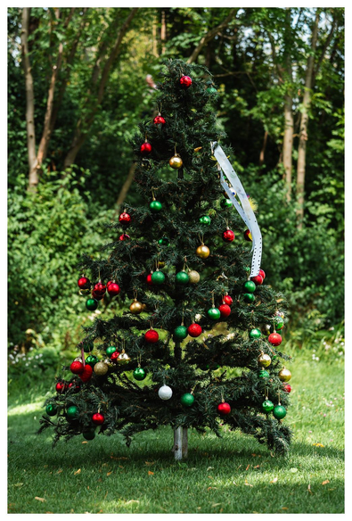 Ein mit grünen, goldenen und roten Kugeln geschmückter (künstlicher) Weihnachtsbaum steht auf einer Wiese in einem Park.
