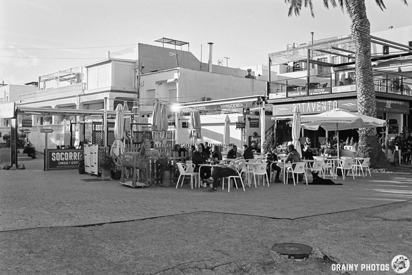 A lively outdoor café scene with tables and chairs, featuring diners under umbrellas. The backdrop includes modern buildings and a palm tree, creating a vibrant atmosphere in a coastal area. Black and white photography highlights the charm of the location.