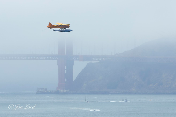 This is a colour photo of a bright yellow seaplane flying across the front of the Golden Gate Bridge on a misty morning with a few boats in the bay below. San Francisco (2017).

The still, light blue waters of the San Francisco Bay rise upwards from the base of the image to a little less than the quarter up level. On the water and about one km away, are a few small boats, including a speed boat heading towards the camera and a sailing boat behind. To the left and above centre is a small bright yellow seaplane inflight from left to right. The aircraft is viewed in profile with it's right side in view and perhaps five hundred metres away. While the fuselage and wings are yellow, the two floats underneath are silver. The plane's registration can be seen on the rear of the fuselage, N123JL. The background is partially obscured by a sea fog. A steep headland descends from the right edge of the photo downwards toward the centre and where it enters the water. There, there is a small building with a small lighthouse on the water's edge. Behind and above the lighthouse is the Golden Gate Bridge. The car deck with it's lattice frame below is partially obscured by the sea fog and extends from the right side, through the centre and over to the left side of the image. Slightly to the left of centre, rising from the base of the headland is the north tower that supports that half of the bridge. The colour, 'international orange' can just be made out. A short distance above the car deck the tower disappears and the tower and sky above are totally obscured by the fog. 