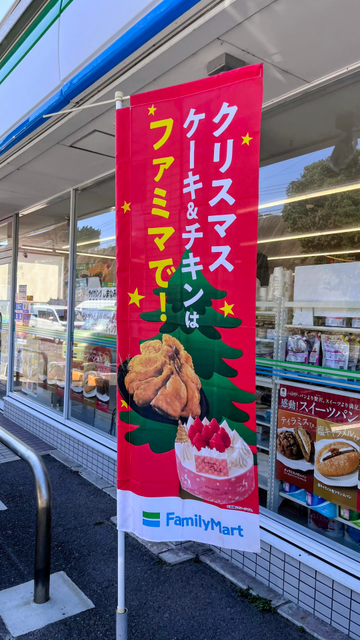 Vor einem FamilyMart Konbini, einem Minimarkt mit Snacks und Erfrischungen, ist hochkant ein roter Aufsteller mit einem grünen Tannenbaum und panierten Hähnchenteilchen, die gugelhupfartig drapiert sind.
Im Schaufenster spiegelt sich der blaue Himmel.
