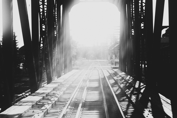 Looking along the tracks of a trestle rail bridge into the setting sun. The glare of the sun washes out a lot of the image in the center with the ridge structure being seen rising on either side. This is a black and white image.