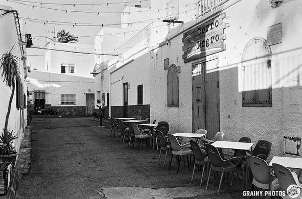 A narrow, sunlit alleyway lined with whitewashed buildings. Empty tables and colorful chairs are set outside, and string lights hang overhead, creating a serene and inviting atmosphere.