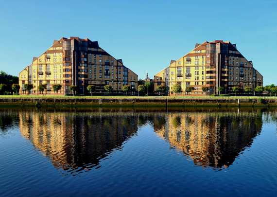 Modern buildings against a clear blue sky reflected in the calm waters in front of them.