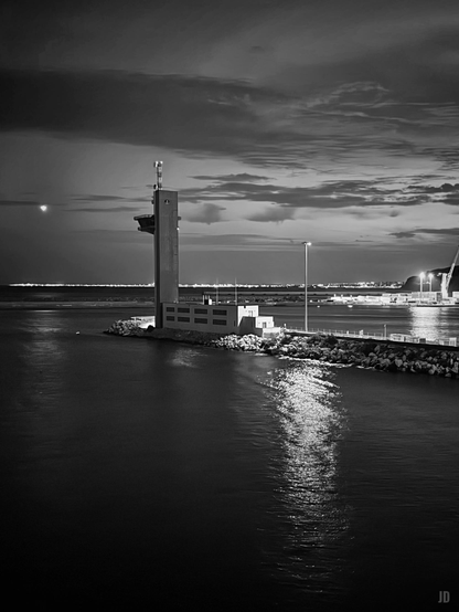 La imagen muestra una escena nocturna en blanco y negro de un puerto. En primer plano, se ve una torre de control o vigilancia situada sobre un espigón rodeado de rocas. El agua refleja la luz, creando un sendero brillante en medio de la oscuridad. Al fondo, las luces de la ciudad y un cielo parcialmente nublado completan el paisaje costero.