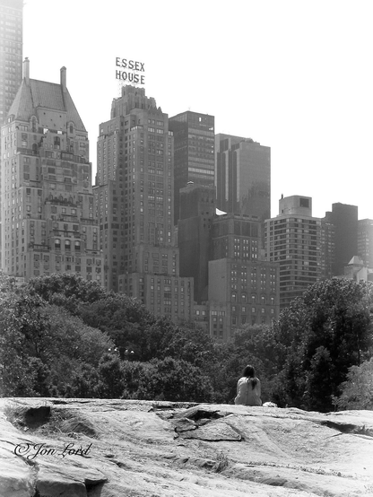 This is a black and white street photo in portrait format of a woman sitting on a large rock with trees in the middle ground and a high-rise cityscape in the background.
Central Park, NYC (2011).

In the foreground is the large rock, spanning the photo from left to right and rising a little less than a quarter up in the image. The light grey rock is fairly smooth with the occasional crack with tufts of grass growing. Below and to the right of centre is our woman, sitting on the rock with her back to the camera and about ten metres away. In the middle ground and beyond the rock is a dense area of dark trees rising to the half way up level. In the background and about a one km away is a wall of high-rise apartment, office blocks and hotels extending diagonally across the photo, the buildings on the left side being the closest and with those on the right getting ever more distant. One high-rise in the centre has the words 'Essex House' in large lettering above the top floor. Above is a bright white sky, devoid of any detail. 