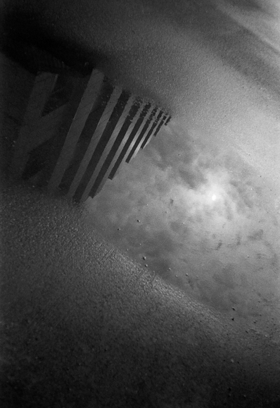 This black-and-white photograph presents a minimalist, almost surreal view of a wet asphalt surface after rain. On the left side of the frame, through the murky, grainy water, the geometric silhouette of a building is reflected — its vertical lines and windows are distorted by the ripples and texture of the puddle. On the right, there is a blurred, bright patch reflecting the overcast sky. The graininess and high contrast make this frame graphic, turning an ordinary puddle into a mirror in which the inverted city looks mysterious and detached.