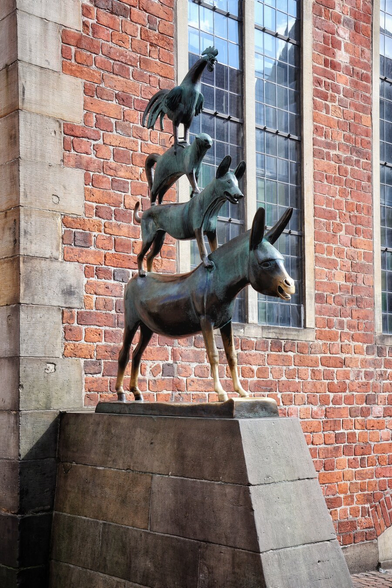 The bronze statue of the Town Musicians of Bremen by Gerhard Marks depicts four animals in a vertical stack: a rooster perches on a cat, which sits on a dog, which stands on a donkey. The sculpture, with its weathered greenish-brown patina, is mounted on a stone pedestal against the wall of Bremen Town Hall, a historic brick building with tall, narrow leaded-glass windows.