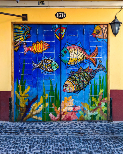 A vibrant painted double door on a yellow building. The door features an underwater scene with several brightly patterned fish swimming above green sea grass and colorful coral at the bottom. In front of the door is a cobblestone street, a black lantern hangs to its right, and an oval plaque with the building number "176" hangs above.