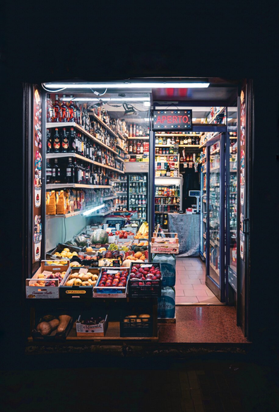This is a photograph of a brightly lit, small corner shop with an open front. The shop is packed with  fresh fruits, vegetables, and bottles. There are shelves in the background which are filled with various beverages and snacks. The inside has fridge display case on the right. The shop has colorful signs. It is framed by dark, shadowed surroundings.