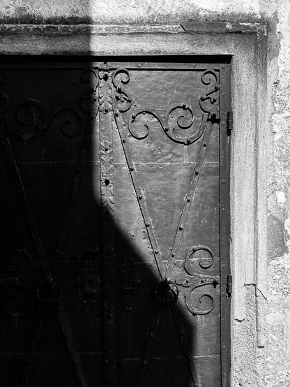 This black and white photo shows an iron door on the side of a church. Hand-forged ornaments and struts made of iron are visible. A dark shadow falls diagonally on the door and bathes it in a mysterious light. It is daytime and the sun is shining.