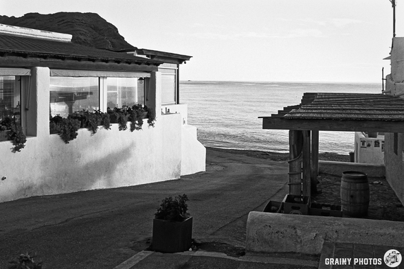 A quiet coastal scene featuring a winding road, a restaurant with flower pots, and a distant view of the ocean under a serene sky. The image is in black and white, capturing a peaceful atmosphere.