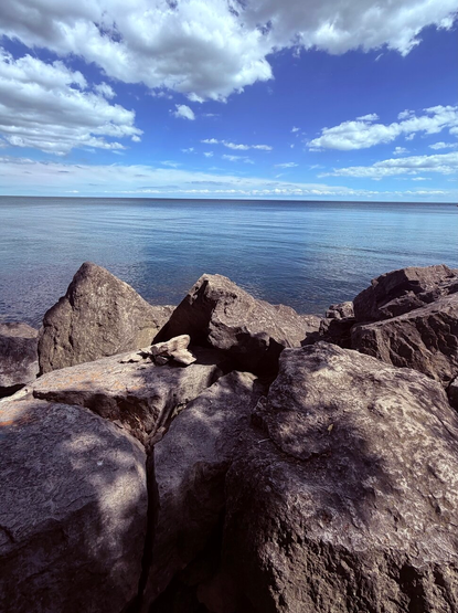 Photograph of the blue sea taken from large grey rocks, under a blue sky with white clouds. The day is sunny and the shadows of a few nearby trees are cast on the rocks, while the white clouds are reflected on the surface of the water.

Photographie de la mer bleue prise à partie de gros rochers gris, sous un ciel bleu avec des nuages blancs. La journée est ensoleillée et l'ombre de quelques arbres tout près est projetée sur les rochers, alors que les nuages blancs sont réfléchis à la surface de l'eau.