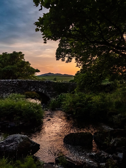 An old stone arch bridge spans a rocky river in the Welsh countryside during sunset. Lush green bushes and overhanging tree branches surround a pool in the river which reflects golden light and deep shadows. In the far distance, rolling fields lead toward a silhouetted mountain range beneath a warm orange and pink sky.