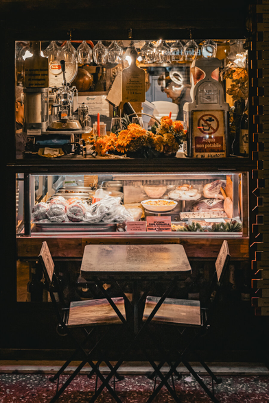 This is a photograph of a dimly lit, rustic restaurant window at night. The window has a glass display case with various meats, including sausages and steaks and a bouquet of orange flowers on top. Above all that there are hanging glasses and on the left there is a coffee machine visible. In the foreground there are rwo chairs with metal legs and a matching table underneath the window.