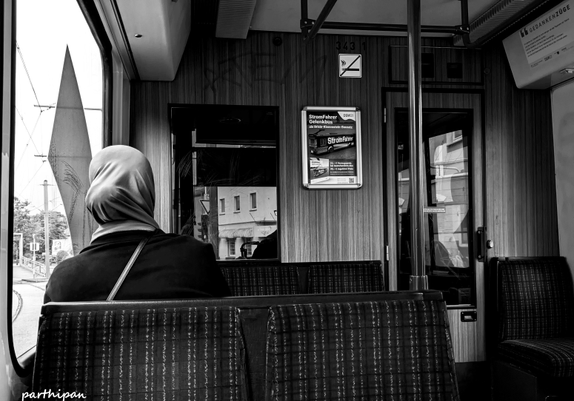A person wearing a headscarf sits alone on a patterned seat in a tram, with their face turned away from the camera; through the windows, city buildings and railway tracks can be seen.

Eine Person mit Kopftuch sitzt allein auf einem gemusterten Sitz in einer Strassenbahn, mit dem Gesicht von der Kamera abgewandt; durch die Fenster sind Stadtgebaeude und Gleise zu sehen.
