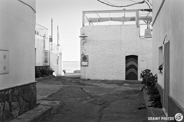 A narrow, winding street bordered by whitewashed buildings leads to a seaside view. A colorful door with stripes stands out against a textured wall, suggesting a tranquil coastal atmosphere in a monochrome setting.