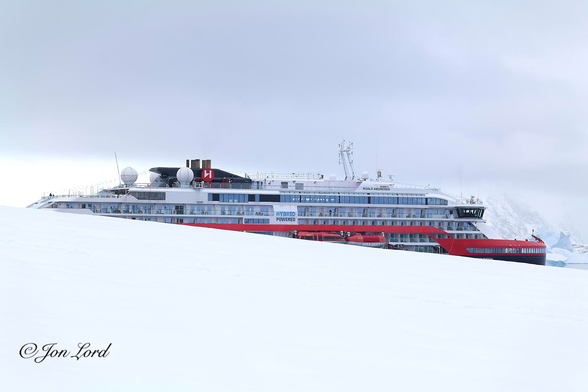 This is a colour photo of an expedition ship with a sloping mass of brilliant white snow in the foreground. Booth Island, Antarctica (2024).

From the centre of the left edge of the photo and sloping gently downwards towards the lower right corner is an expanse of smooth brilliant white snow. Beyond the snow, centred in the image and partially obscured by the snow, is the expedition ship. Viewed in profile, the right side in view, facing to the right and occupying nearly all of the width of the photo. Only a segment of the upper half of the vessel is in view. The ship has a black hull, a thick, bright red stripe running along the length of the ship and a white superstructure above. Two large covered lifeboats with white hulls and 'international orange' coloured (day glow) cabins above, hang from davits amidships. Aft and above the lifeboats are the words "Hybrid Powered" in large blue letters. On the lifeboat deck and the two decks above are rows of cabins, many with balconies. Less than a handful of hardy passengers can be seen scattered about the upper and aft deck. Aft of the bridge is the ship's name "Roald Amundsen" in large black letters. Above and aft of the upper deck is a raked black funnel with a red roundel with a large white letter H, the shipping lines logo and with numerous black vertical exhaust pipes extending upwards. Forward of the ship's bow in the lower right corner is a hint of the frigid waters of the Antarctic with some large, white and turquoise ice bergs with snow and rock covered mountains beyond. The sky is obscured by a low ceiling of cloud. 

The ship is the 140m long, MS Roald Amundsen. 

