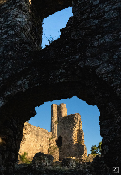 Color photo of the sunlit tower of a 12th century castle in Catalonia Spain viewed through a shadowed rough doorway opening in a ruined wall, with a window opening above it showing blue sky. 