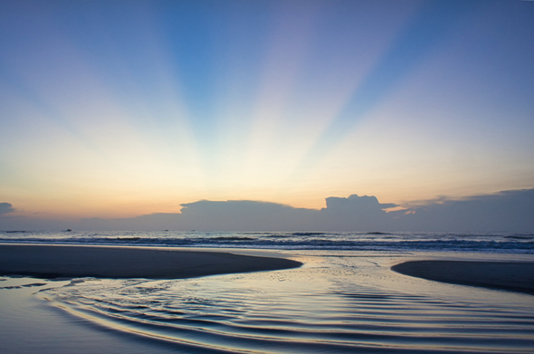 Sunrise over a serene beach, vibrant rays fan out behind distant clouds, reflecting on calm water with gentle ripples, creating a peaceful atmosphere.