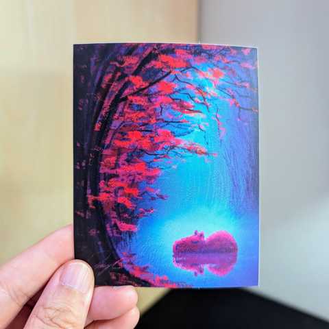Photo of a left hand holding a rectangle sticker. The sticker is a matte painting of a capybara resting under fall leaves.