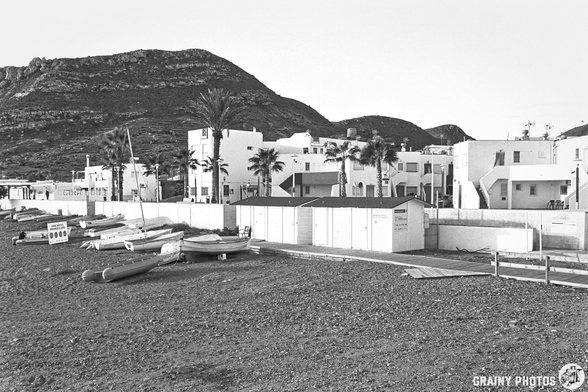 A serene beach scene featuring white buildings and palm trees set against a mountainous backdrop. Several boats are lined up on the sandy shore, giving a tranquil coastal vibe. The image is in black and white, emphasising the calming atmosphere.