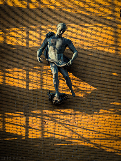 A bronze statue of a young, athletic male figure. The statue is mounted on a large, uniform wall made of yellowish bricks.

The scene is illuminated by warm, golden-hour sunlight casting strong, graphic shadows. A prominent, grid-like geometric shadow from scaffolding, stretches across the wall. 

The statue is mounted on the façade of Studio-I. The sculpture is made by Pieter Starreveld (1911–1989).