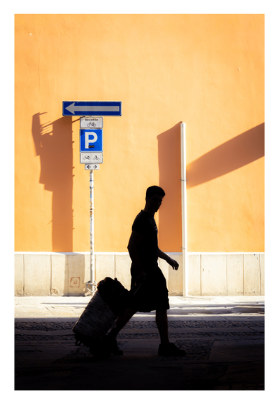 This is a photograph of the dark silhouette of a person carrying a suitcase behind them. They are walking towards the right hand side of the frame. Below them them cobbled street is also dark and shadowed. Behind them on the other side of the street there is a bright yellow wall with bricks lining the part closest to the ground. On the left of the silhouette there is a blue "P" parking sign and on the right there is a tube.