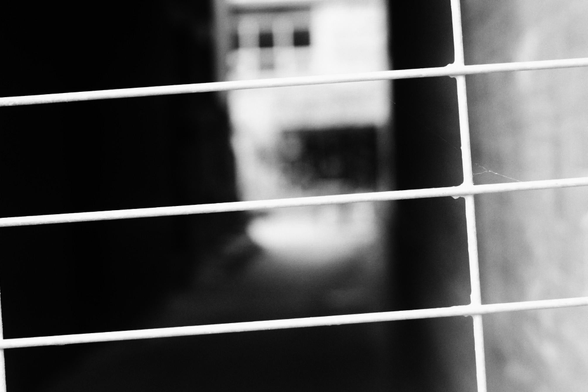 Standing behind a wire fence looking towards the blurry out of focus view down a narrow alley. The fence can be seen in detail in this black and white image.