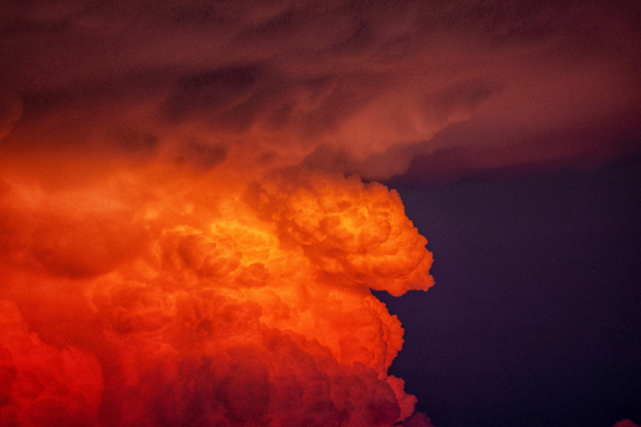 Das Bild zeigt eine gewaltige, dramatische Wolkenformation, die in ein intensives Licht getaucht ist und fast wie ein loderndes Feuer am Himmel wirkt. Im Zentrum dominiert eine massive, bauschige Cumulonimbus-Wolke, deren Struktur an einen emporsteigenden Atompilz oder brodelndes Magma erinnert. Diese zentrale Wolkenmasse leuchtet in einem extrem kräftigen Orange und glühenden Gelb, was auf eine tiefstehende Sonne während eines Sonnenuntergangs hindeutet. Die Oberfläche dieser Wolke ist stark zerklüftet und weist unzählige Schattierungen und Texturen auf, die ihr ein plastisches, dreidimensionales Aussehen verleihen. Im oberen Teil des Bildes gehen diese feurigen Farben in dunklere, violette und tiefrote Töne über, wobei die Wolken dort flacher und flächiger wirken. Rechts im Bild wird die leuchtende Formation durch einen fast schwarzen, tiefvioletten Bereich des Himmels begrenzt, was den Kontrast zur glühenden Wolke massiv verstärkt und die Szenerie besonders theatralisch und fast schon apokalyptisch erscheinen lässt.