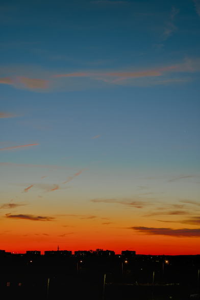 Das Foto zeigt einen farbenprächtigen Sonnenuntergang über einer Stadt.

Der Himmel ist in warmen Orange-, Gelb- und Rottönen gefärbt, die nach oben hin in Blautöne und Lila übergehen. Am Horizont sind die Silhouetten von Gebäuden und Bäumen als dunkle Konturen zu erkennen. Einige Wolken reflektieren die Farben des Sonnenuntergangs.
 --- 
The photo shows a colorful sunset over a city.

The sky is tinged with warm shades of orange, yellow, and red, which fade into shades of blue and purple toward the top. On the horizon, the silhouettes of buildings and trees are visible as dark outlines. Some clouds reflect the colors of the sunset.