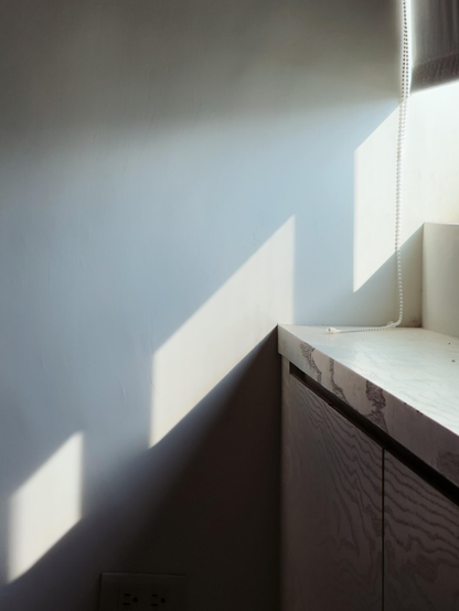 Sunlight casting geometric shadows on a plain white wall near a window with a beaded chain and a wooden surface.