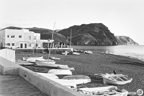 A serene black and white image of a pebbled beach dotted with boats, a coastal building with outdoor seating nearby, and a dramatic rocky landscape in the background, reflecting a tranquil seaside atmosphere.