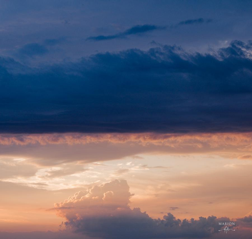 Man sieht einen Abendhimmel die obere Hälfte ist geprägt von intensiven blau und dunklen blauen Wolklen, unterhalb an der blauen Wolkenkante zeigt sich ein schmales Band von rosa Schäfchenwolken, die dann in gold und orangen wolken übergehen, Mittig unten zeigt sich eine dicke Wattewolke in graugold.