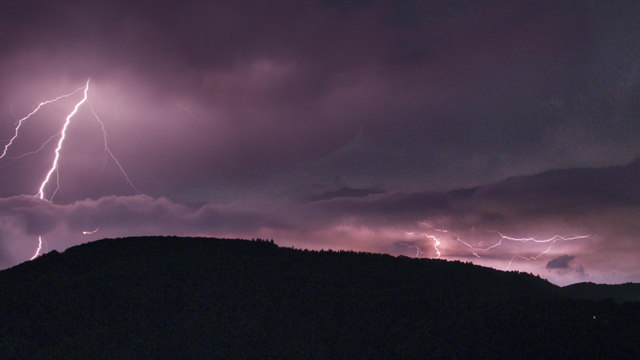 Nachtfoto einer dunklen Hügelspitze, über der im grau-violett-lila-farbenen Himmel mehrere Blitze zu sehen sind, die die dunklen Wolken drumherum jeweils etwas aufhellen.