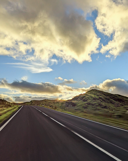 A scenic, two-lane asphalt road in the Welsh countryside with a large green hill to the right and vibrant clouds catching the golden hour light.