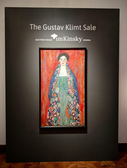 Ein Bild des Künstlers Gustav Klimt, das den Titel "Fräulein Lieser" trägt. Es zeigt eine junge Frau vor rotem Hintergrund. Der Umhang ist mit Ornamenten verziert. Ihr Kleid ist in einem hellen Grün gehalten.